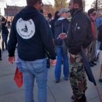 Puget Sound John Brown Gun Club members chat. Both are white men; one wear a JBGC sweatshirt and has a red bandana hanging from his pocket. He wear a cammo-patterned yarmulke. The other smokes a cigarette, wearing a black bandana around his lower face/neck, and cammo pants.
